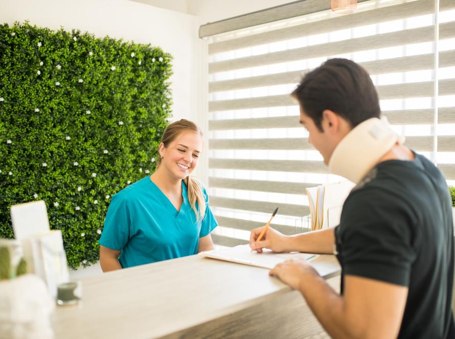 physical-therapist-looking-male-athlete-with-cervical-collar-filling-medical-form-reception-counter-hospital