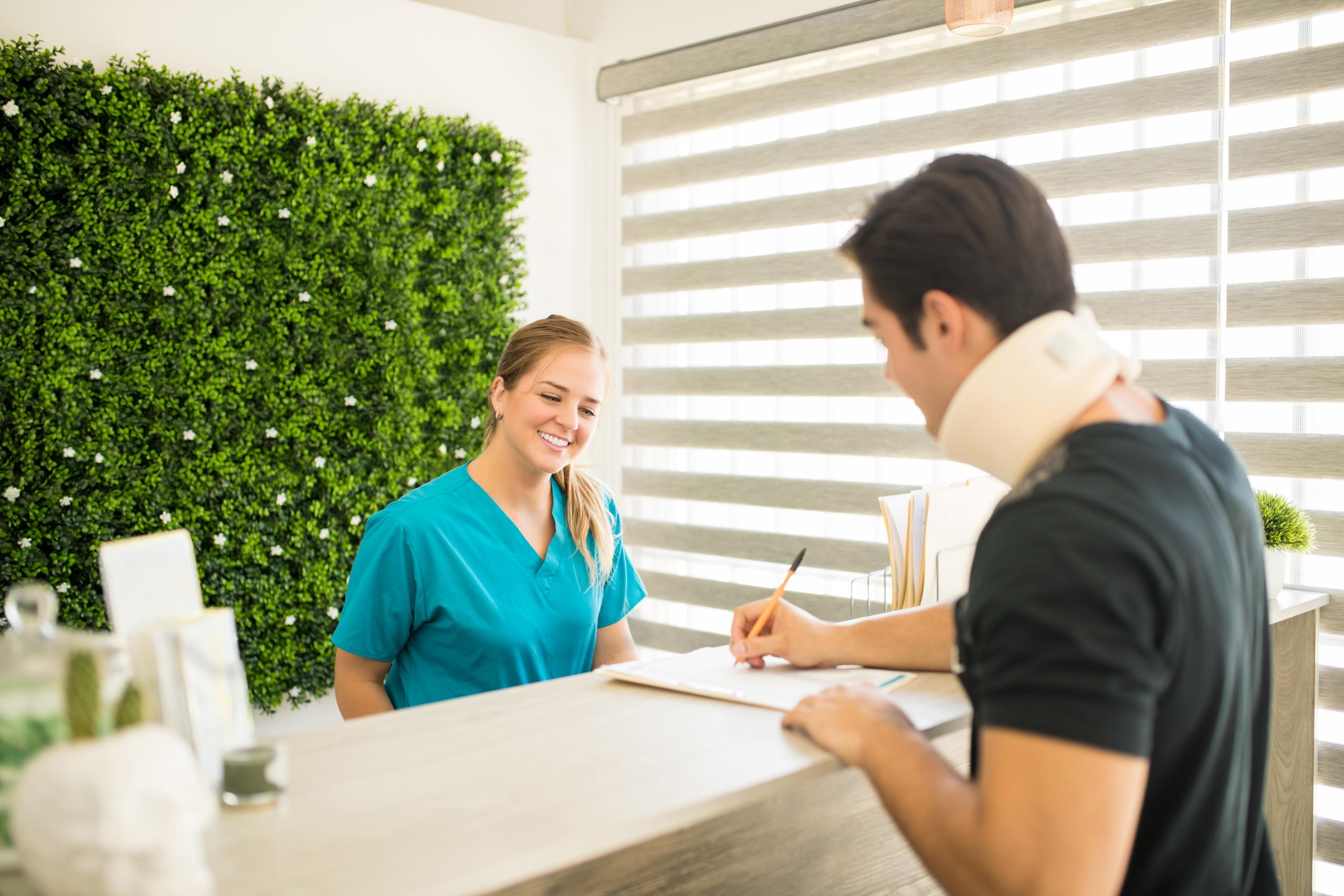 physical-therapist-looking-male-athlete-with-cervical-collar-filling-medical-form-reception-counter-hospital